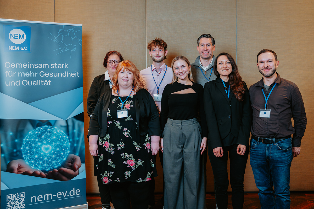 Group of seven people standing indoors in front of a wood-paneled wall, next to a blue banner with text about health and quality from NEM e.V.