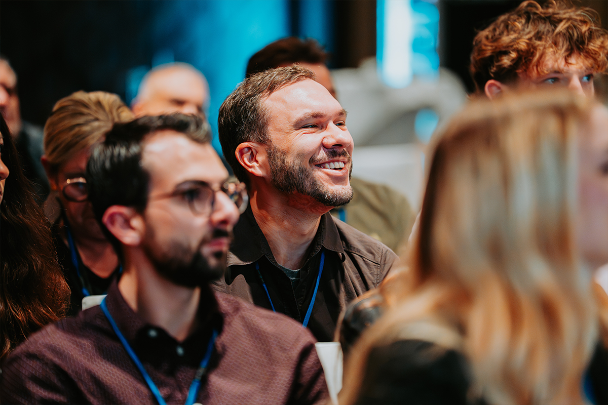 Smiling man with a beard attending an indoor event with other people seated around him.