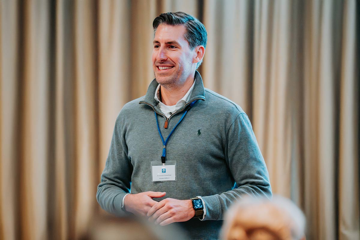 Smiling man in a grey quarter-zip sweater with a name badge and smartwatch, standing against a beige curtain background.